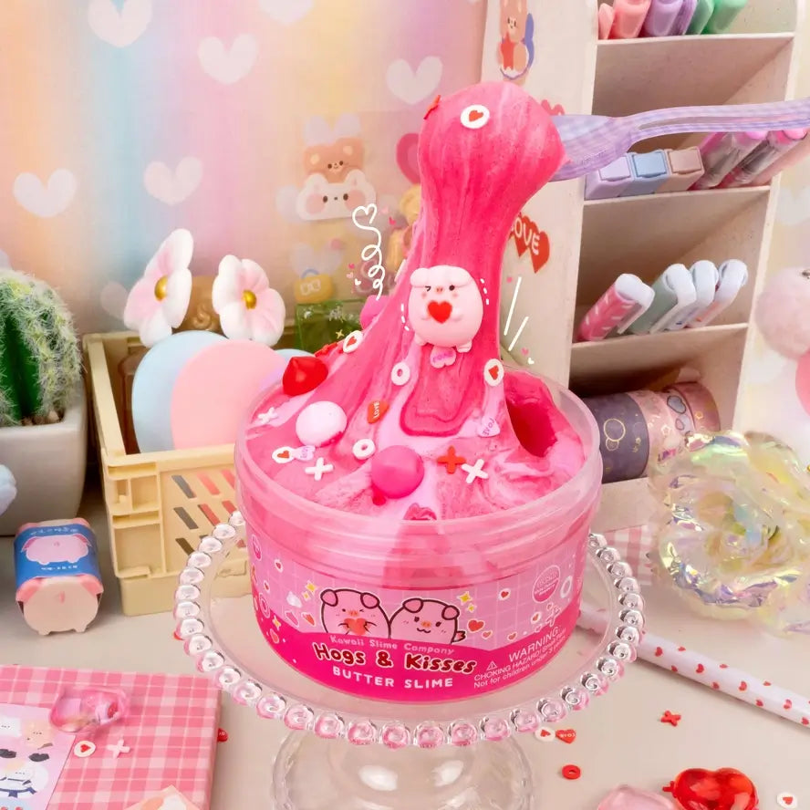 A plastic spoon holds up the pink slime as a pig holding a heart rides the "wave" down to the container for Hogs & Kisses Butter Slime.