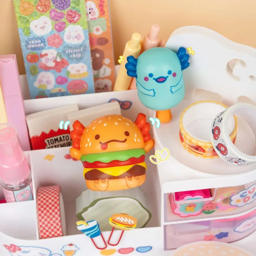 The hamburger and popsicle Snaxolotl sit on a Kawaii display of office supplies.