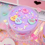 A purple container of fluff slime covered in toy Conversation Hearts, multicolored hearts, and x's and o's.
