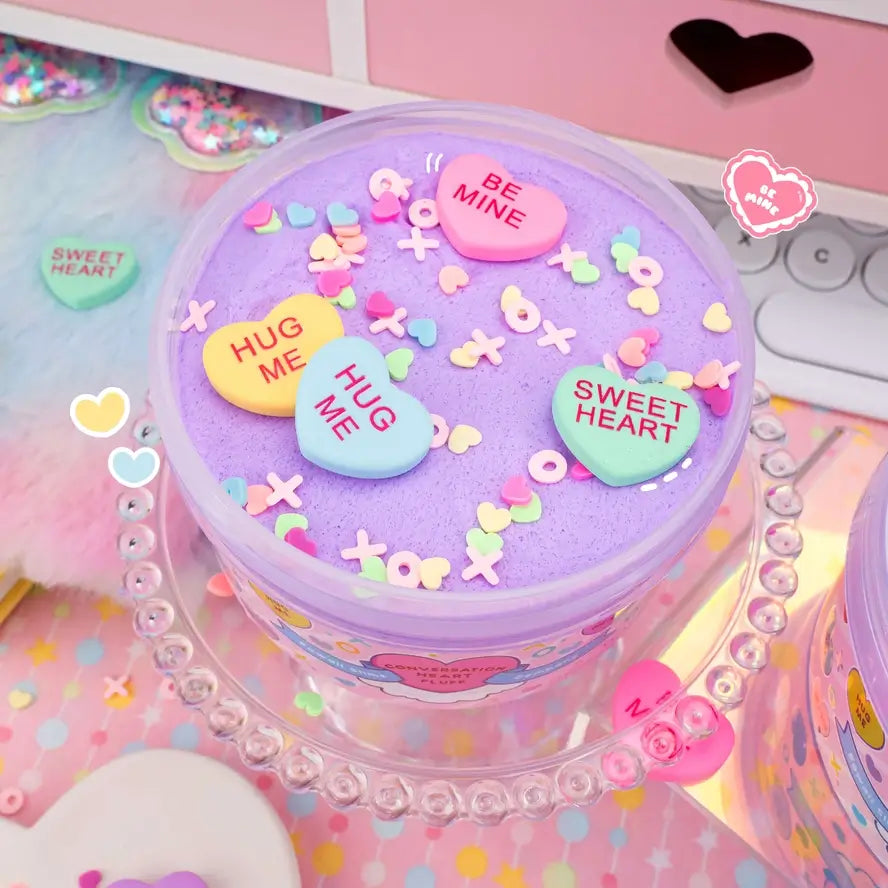 A purple container of fluff slime covered in toy Conversation Hearts, multicolored hearts, and x's and o's.