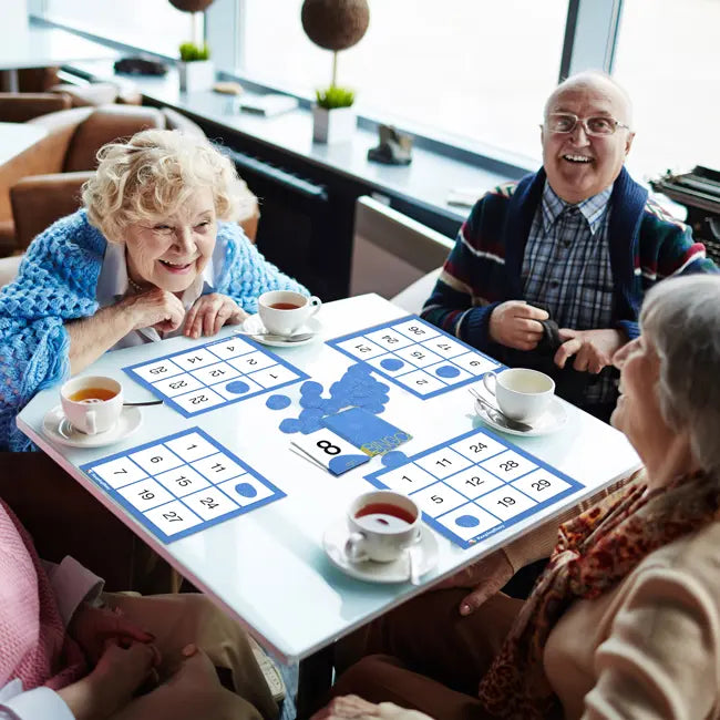 Four seniors sit around a recreation table with cups of tea; they are laughing and playing a game of Easy Bingo.