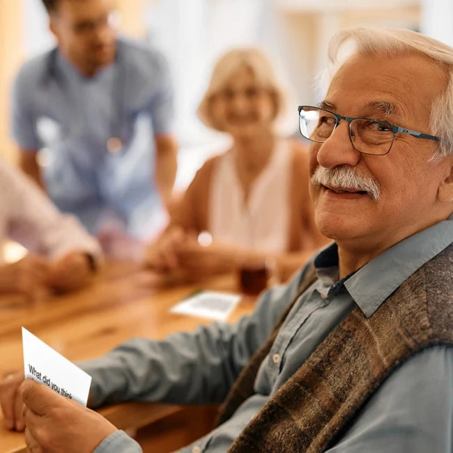 An older man holds a conversation card at a table with a woman and a nurse in the background.