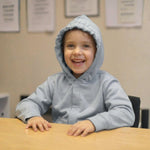 A child smiles at a desk with the hood up on their Grey Kids QuietSense™ Sound Reducing Sensory Hoodie.