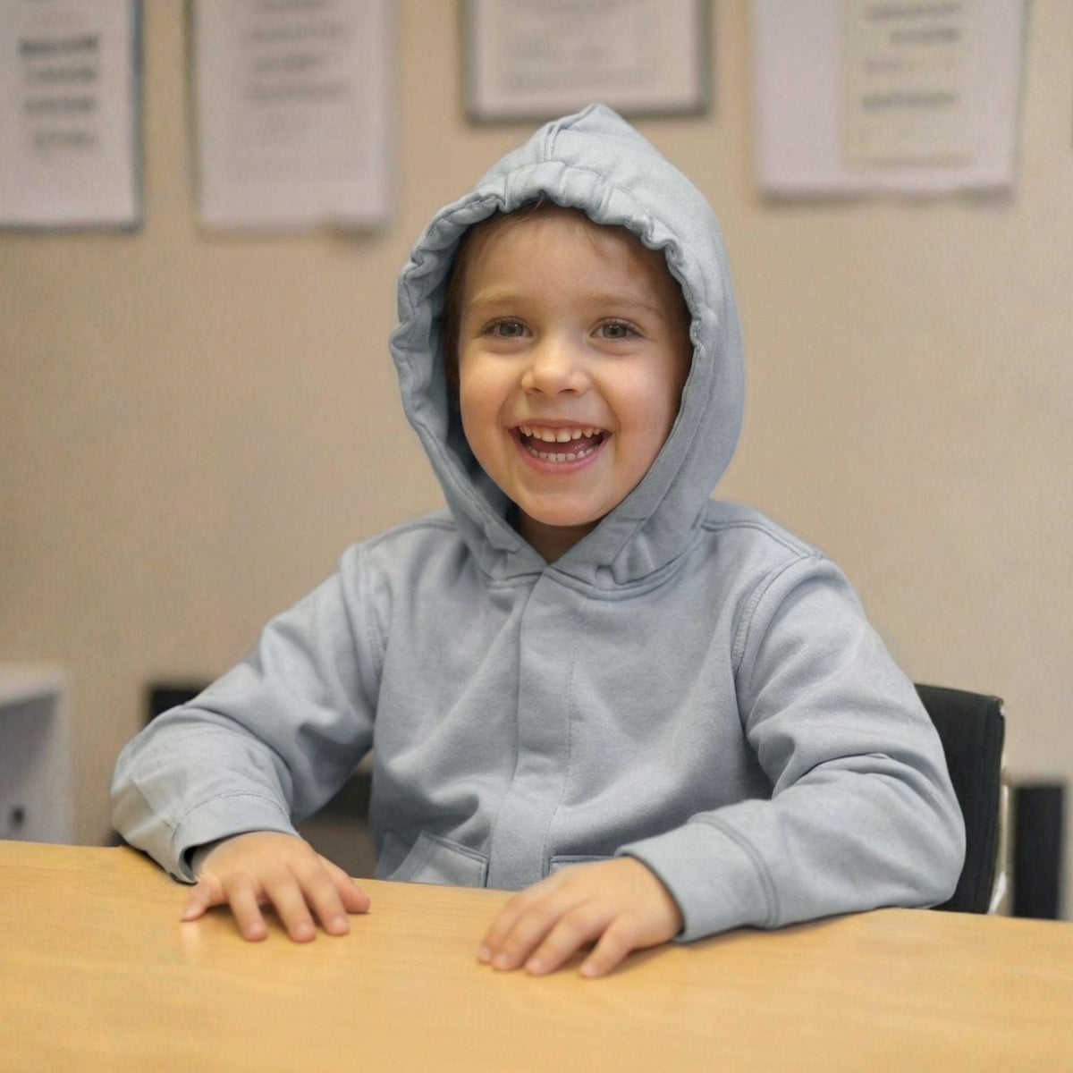 A child smiles at a desk with the hood up on their Grey Kids QuietSense™ Sound Reducing Sensory Hoodie.