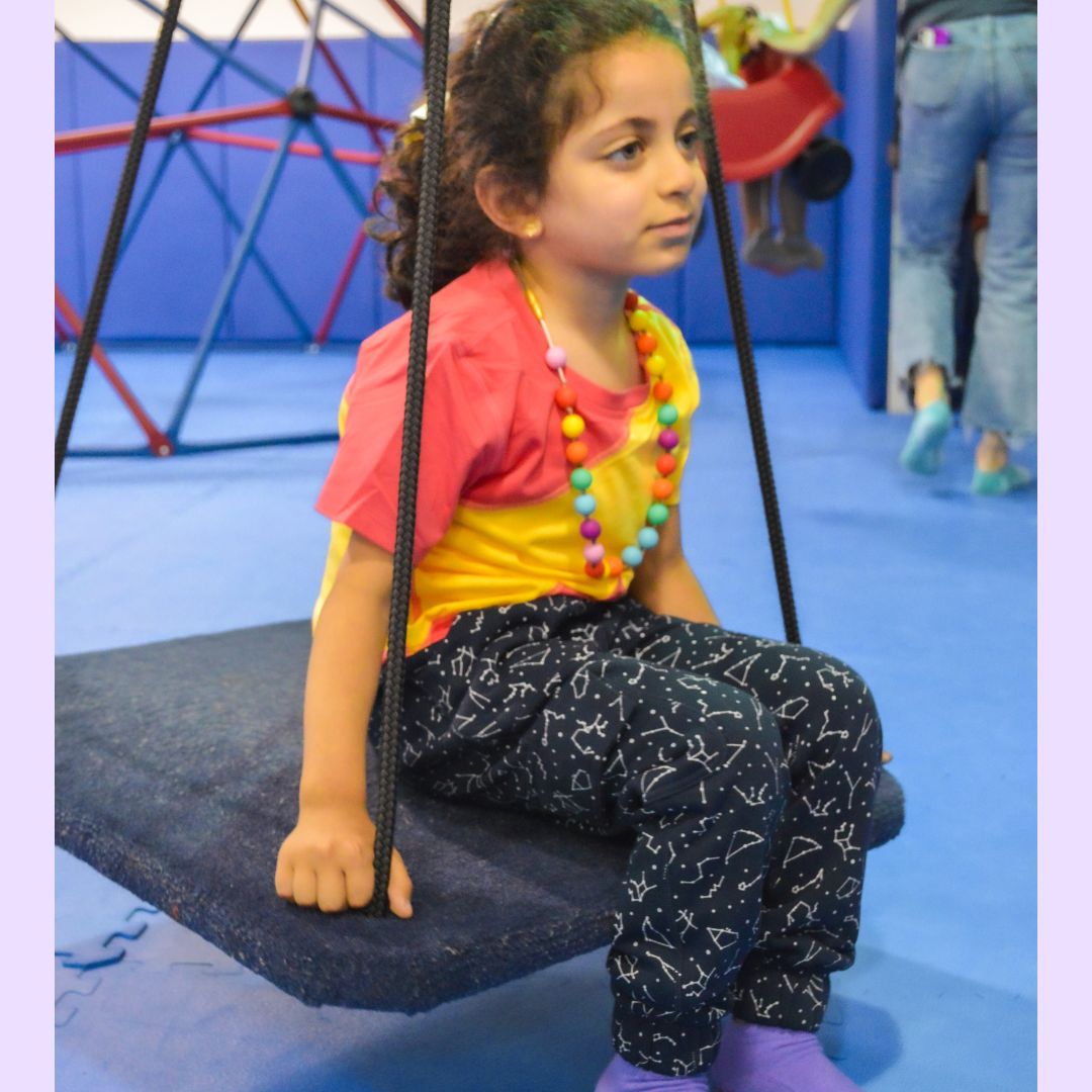 A child sits on a carpeted sensory swing wearing the Contellation Print Kids Sensory Friendly Fidget Jogger.
