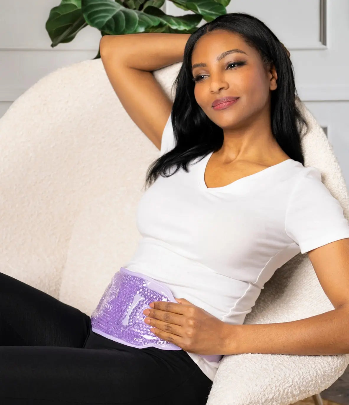 A woman holds the purple Ice & Easy Hot & Cold Body Wrap over her abdomen and leans back in a chair.