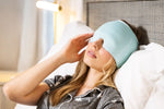 A woman lays back on a pillow in bed while wearing the teal Out Cold Temp-Smart Migraine Cap.