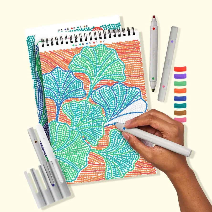 A hand filled in the color blue on a letter on a picture of ginko leaves in the Lifelines FLowArt Dot-by-Letter Botanicals Pad.