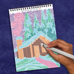 A hand fills in the color matching the letters on this FlowArt scene of a cabin covered in snow in the woods.