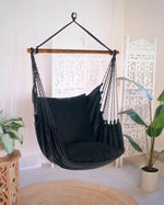 The Black Boho Hammock Chair Swing hangs in a living room decorated with plants.
