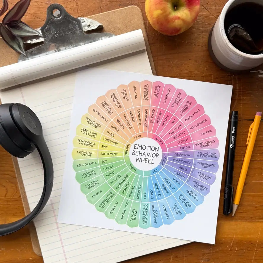 The colorful Fidget Proof Emotion Behavior Wheel sits on top of a lined notebook, next to some writing utensils and a pair of headphones.