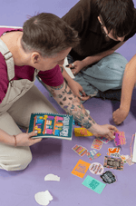 Two people sit on a floor with a Sticker Library and look through a pile of stickers.