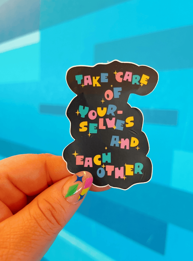 A hand with a very cute manicure holds up the Take Care of Yourselves and Each Other Sticker.