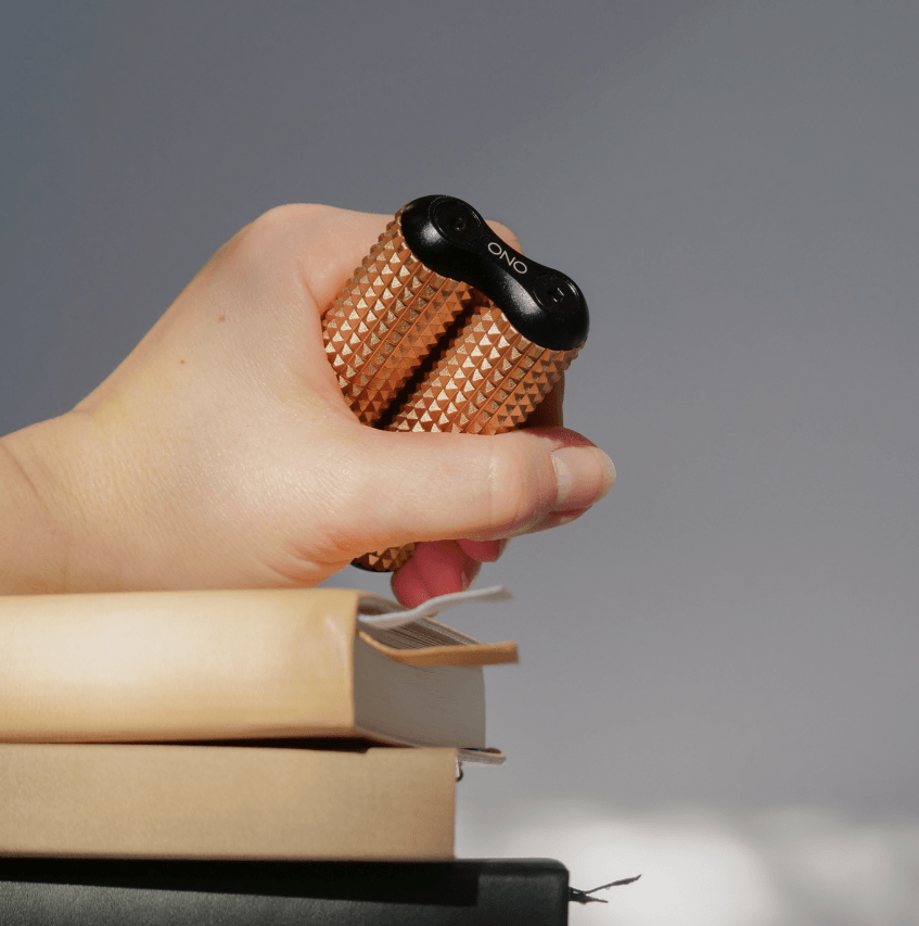 A hand holds the Peach ONO Aluminum Acupressure Roller over a pile of books.