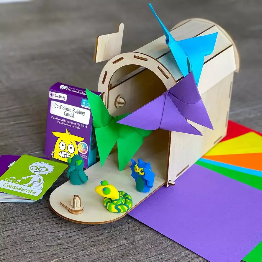 A small wooden box is surrounded by little clay figures, folded paper butterflies, and Confidence Building Cards.