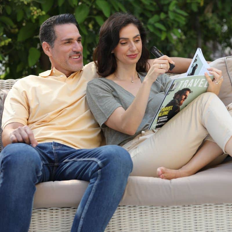 Two people sit on a couch next to each other; one is using the OrCam Read 3 to look at a magazine.