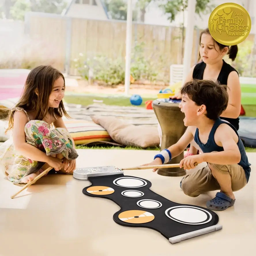 Two children sit next to the Roll-Up Electronic Drum Kit. Each holds one stick and hover over the kit rolled out on the floor, while a third child holds a small drum.