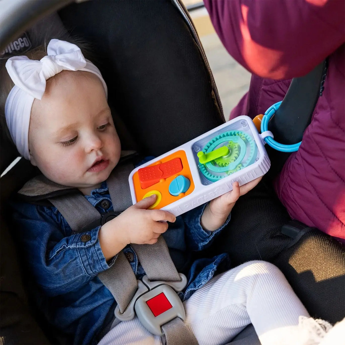 A baby holds the PlayTab Go Travel Sensory Activity Board while strapped into a car seat.