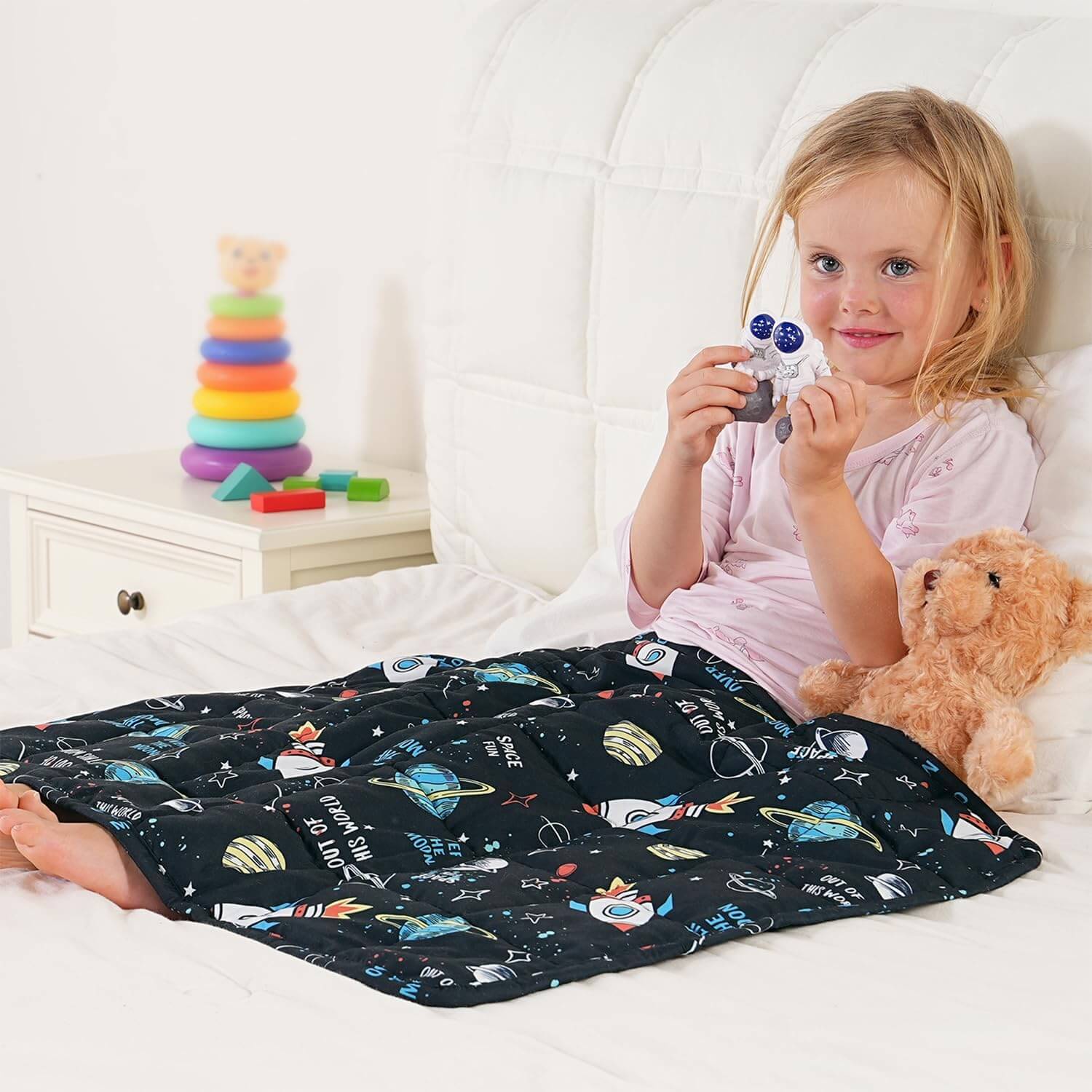 A child holds up two astronaut toys while laying in bed, covered by the Rocket Weighted Cooling Lap Pad 2 lb.
