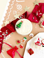 Several of the charms from the Santa's Cottage KidDough Kit are spread out on a table next to an open container of the Cookies & Cream KidDough.