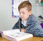 A child sits over an open book at a desk while wearing the Sensory Weighted Shoulder Wrap.