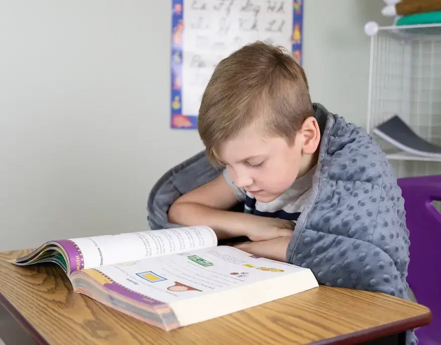 A child wears the Sensory Weighted Shoulder Wrap while learning over an open book at a desk.