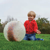 A child with light skin tone and short blonde hair is kneeling on grass and pushing the Constellation Sensory Ball.