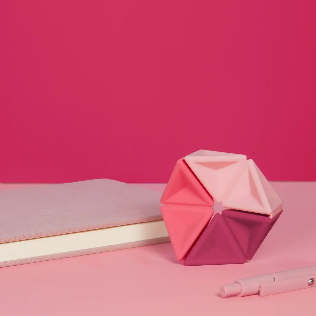 The Speks Magenta Pivot Silicone Flexagon next to a pink notebook.