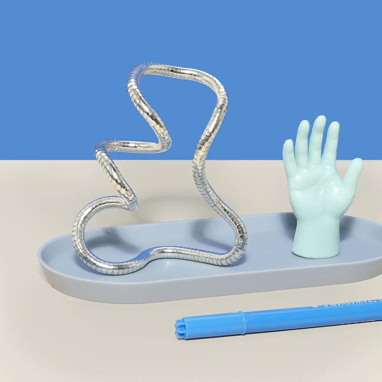The molded silver Speks Strand Moldable Fidget Rope on a grey platform next to a tiny toy hand.