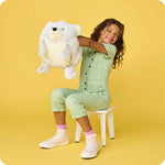A child sits on a stool and holds the  Supersized Bear Hand Warmies up by its side pockets.
