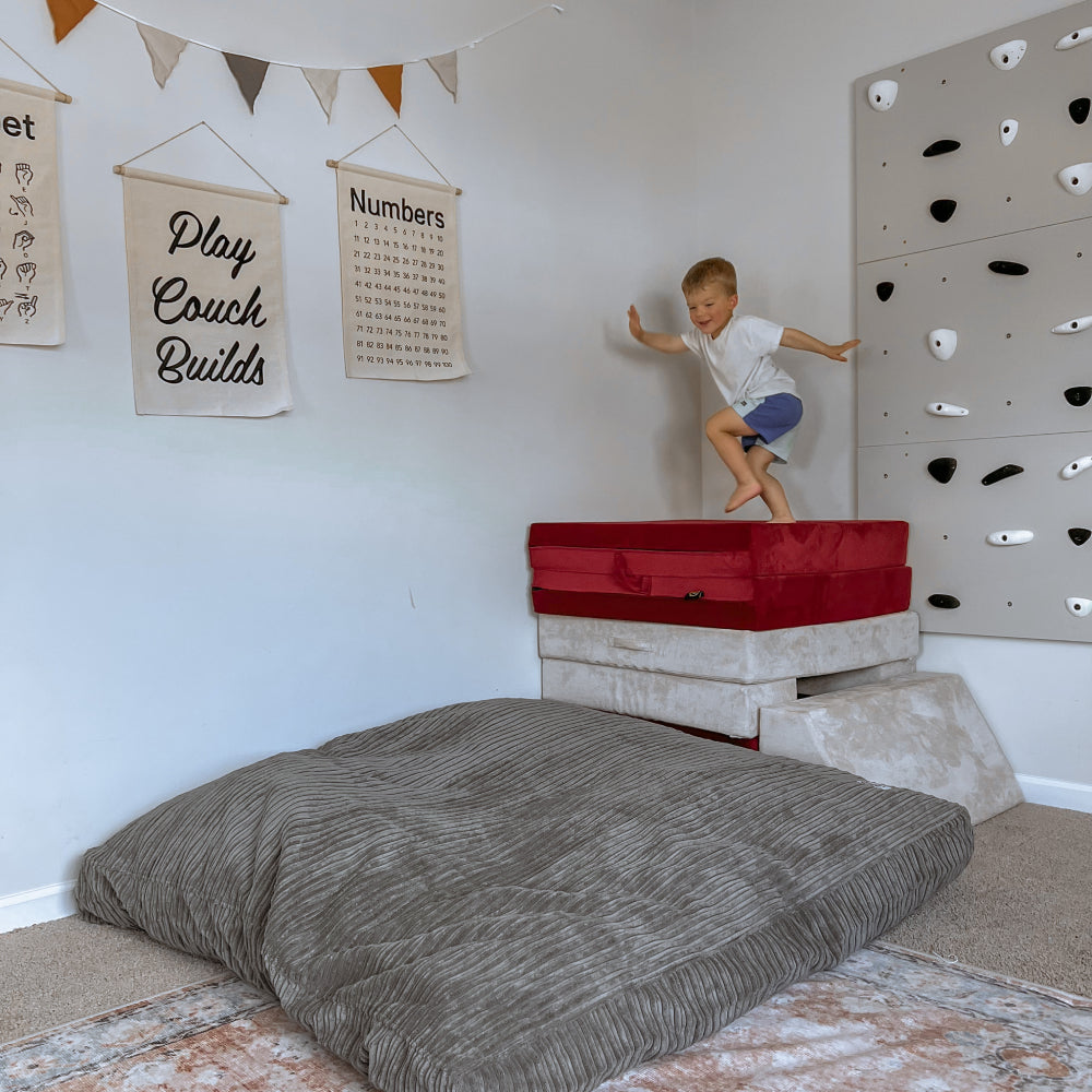 A child is positioned to jump off of the soft tower and onto the gray Terry Corduroy Sensory Crash Pad below.