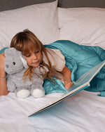 A child cuddles with a stuffy while reading with the Aqua Mini Baloo Weighted Blanket.