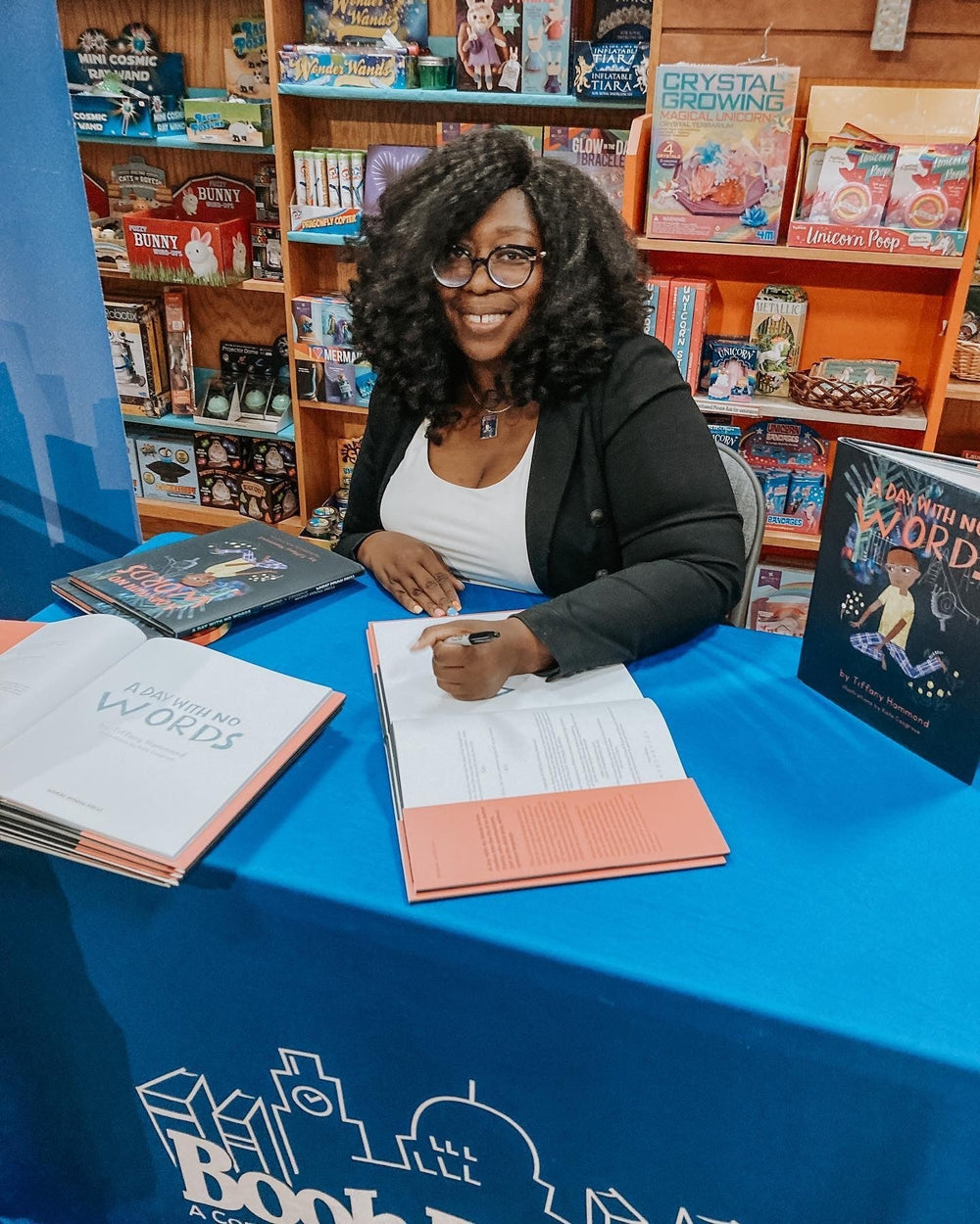 A dark skinned woman with glasses and curly, shoulder length hair sits at a table during a book signing event with a book open and a pen perched above a page.