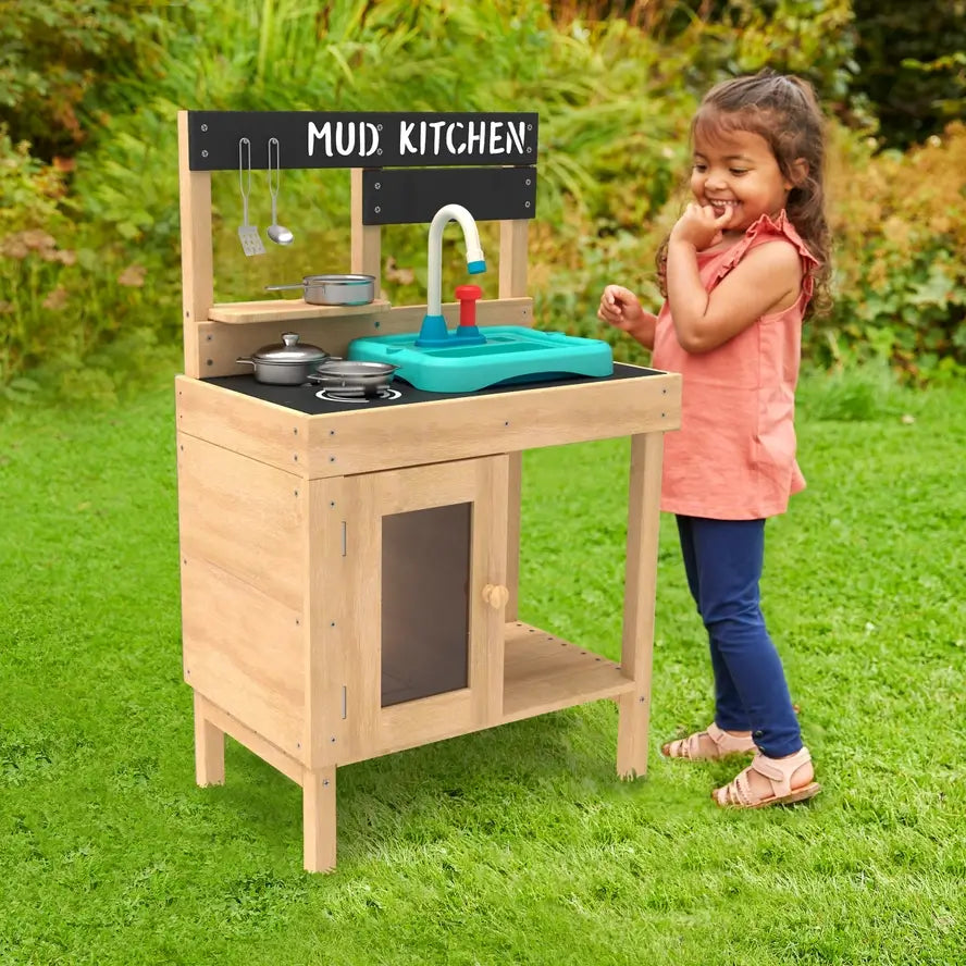 A child stands on backyard grass while smiling at the TP Toys Splash and Play Early Fun Wooden and Kitchen Play Set.