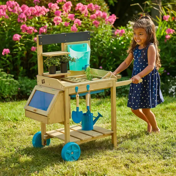 A child pushes the TP Toys Wooden Explore Potting Bench Outdoor Playset over grass in a yard.