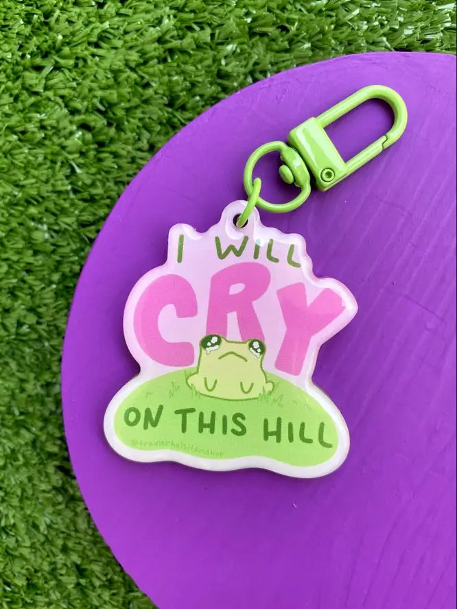 The "I Will Cry on This Hill" keychain on a purple surface.