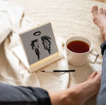 A pair of angel wings and a halo painted on the White Mini Buddha Water Drawing Board on a bed next to a cup of tea.