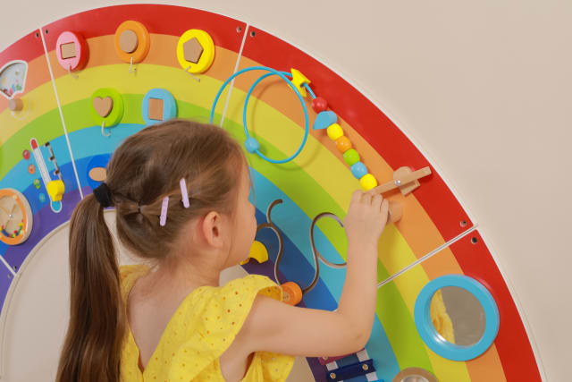 A child with light skin tone and long brown hair plays with the wooden beads on the Rainbow Activity Wall Panels.