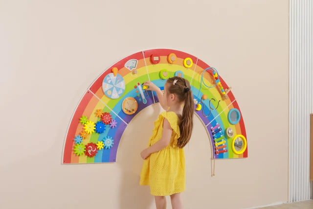A child with light skin tone and long brown hair plays with the thermometer on the Rainbow Activity Wall Panels.