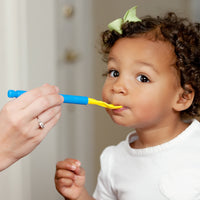 A child with medium skin tone and short curly hair is looking up and to the side as an adult hand holds a Z-Vibe with the yellow Smooth Spoon Tip in the child's mouth.