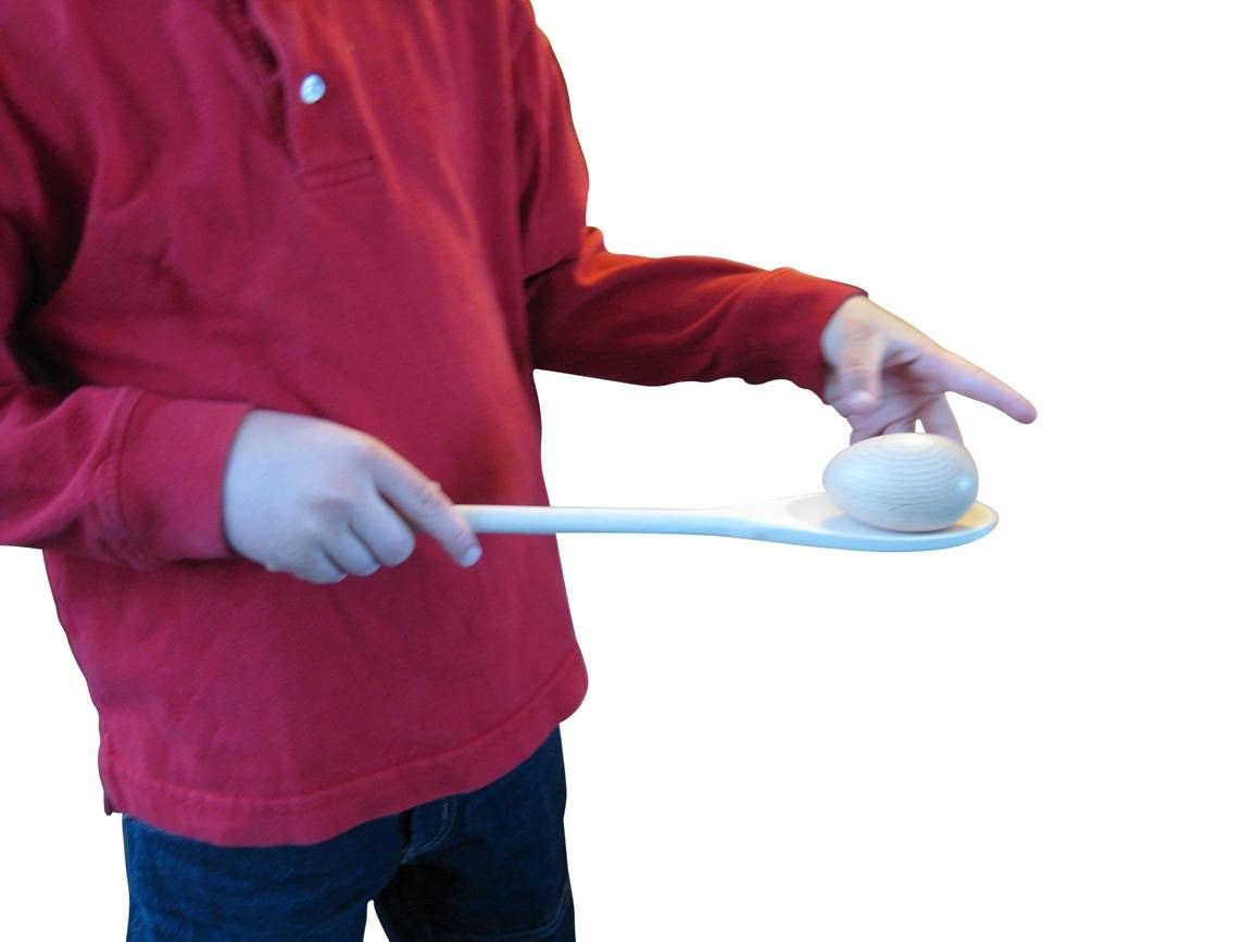 A child's torso holds a wooden spoon with one hand and the other hand is hovering over the egg sitting at its tip.