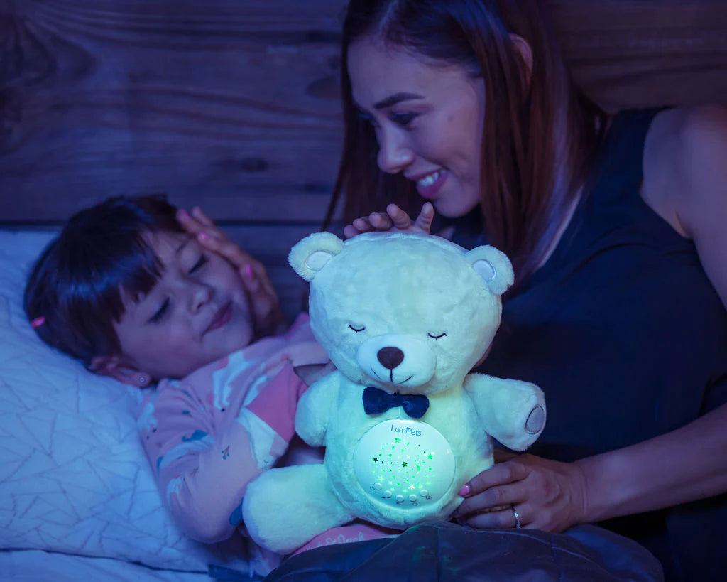 A person with medium dark skin tone and long brown hair smiles while holding a Lumipets Sound Soother Bear next to a child who is lying on a bed. The child has medium skin tone and brown bangs.