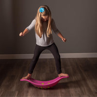 A child with light skin tone and long blonde hair is standing with either foot over the edges of the pink Teeter Popper. They are looking down and balancing.