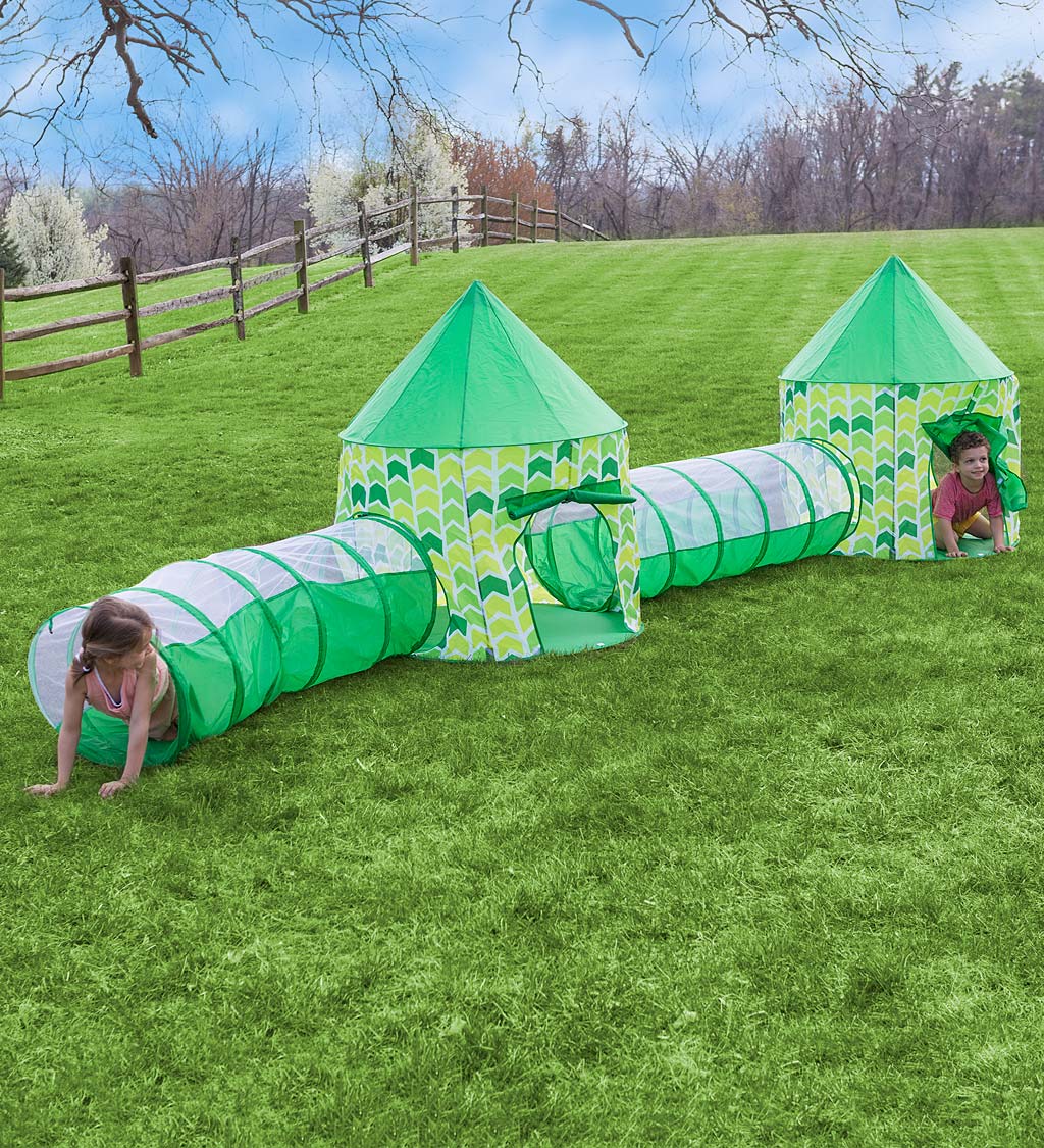 The set of four tents and tunnels is outside on the grass. One child with light skin tone and light brown hair pulled back is coming out of a tunnel and propping themselves up with their arms. Another child with light skin and short brown hair is at the far end of the structure, peaking their head out of a tent while on their hands and knees.