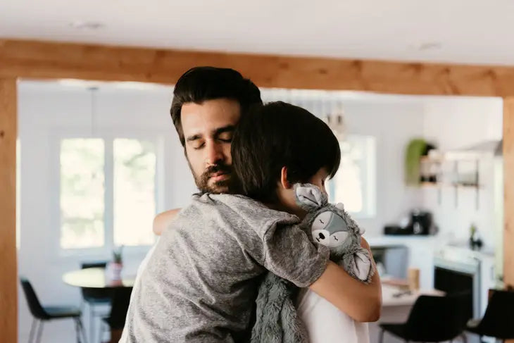 A child with light skin tone and black hair is holding the Slumberkins Slate Ibex Snuggler and hugging an adult with light skin tone and black hair and a beard.