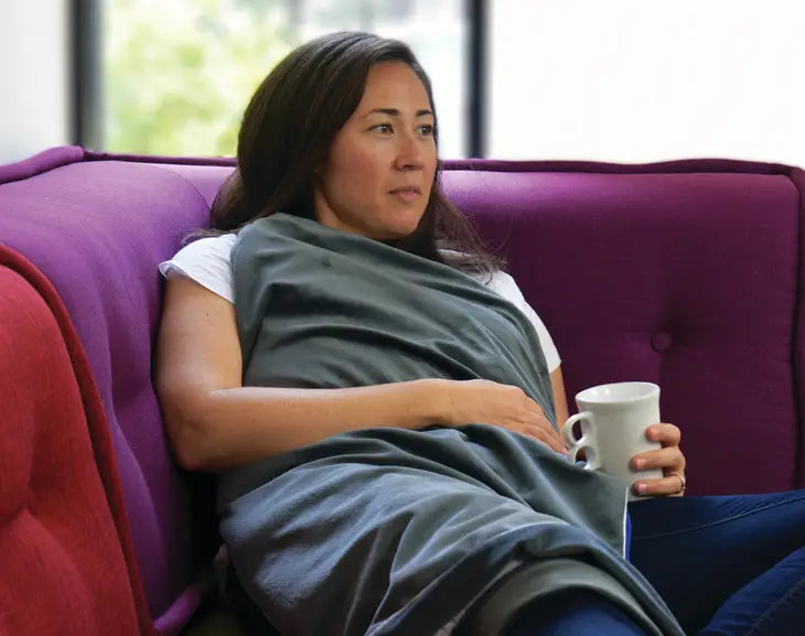 A person with medium light skin tone and long bown hair lays back on a couch with the Ms Bliss Aroma Blaket on their chest and legs. They have one arm folded over the blanket, and the other is propping up a mug on their leg.