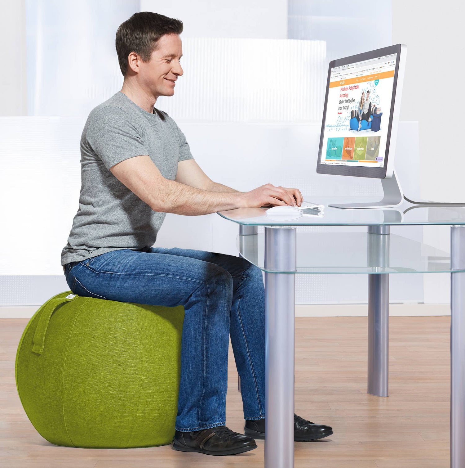 A person with light skin tone sits on a Green YogaBo at a desk with a computer. They are smiling and looking down at the keyboard.