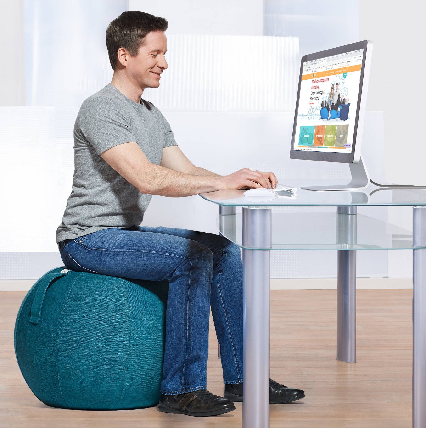 A person with light skin tone and short brown hair sits at a desk on the Turquoise YogaBo. They are smiling and looking down at the keyboard in front of them.