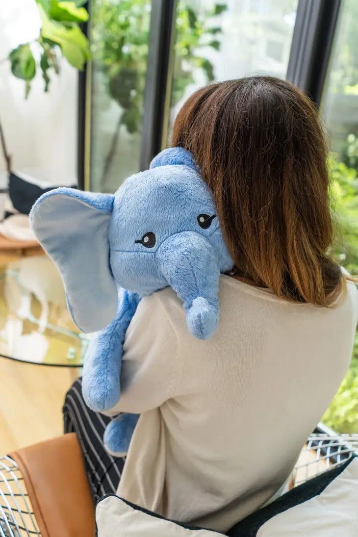 A person with long brown hair is holding the Elephant Hugimals Weighted Stuffed Animal over their left shoulder.