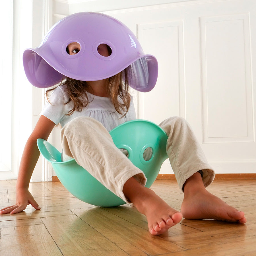A child with light skin tone and shoulder length brown hair sits in a mint Bilibo and balances a lilac Bilibo on their head. Their eyes are peaking out through the two holes on the seat.